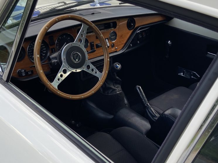 1973 Triumph GT6 interior shot showing wooden steering wheel, dash and gauges from the driver's side window